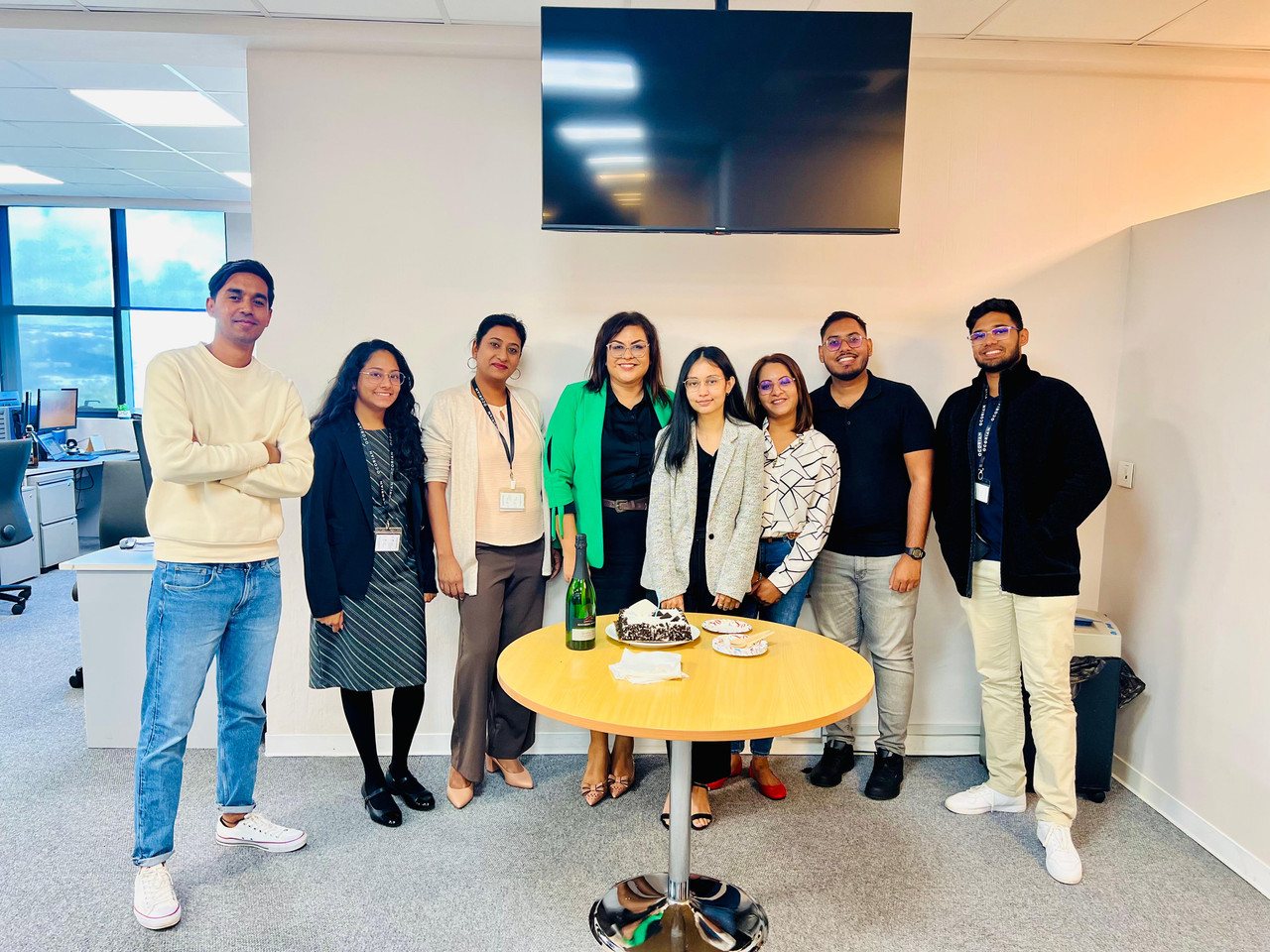 Group of Ocorian colleagues gathered around a small round table holding a cake and a bottle of sparkling wine, in an open-plan office with several wearing lanyards printed with the Ocorian name
