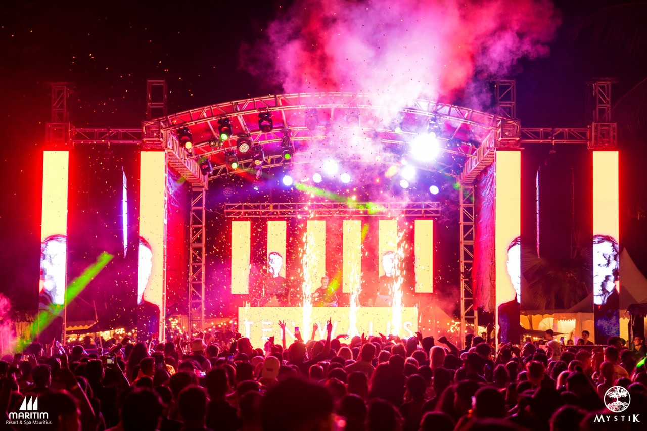 Festival stage at night with vertical LED screens, spark machines and confetti over a dense crowd