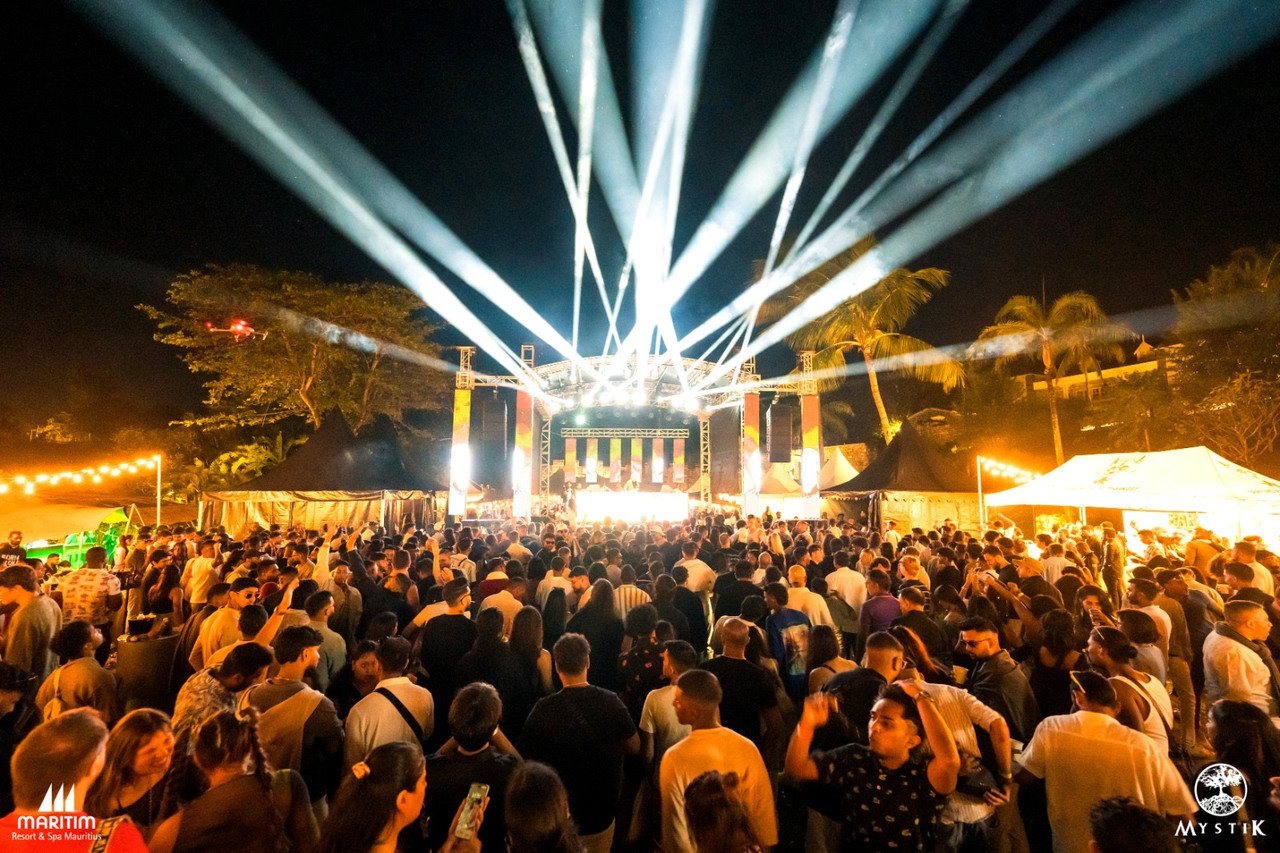 Night view of the main festival stage from behind the crowd, with searchlight beams and a drone in the sky