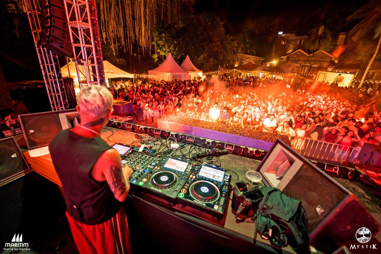 View from the DJ booth over a packed outdoor festival crowd lit in red and orange at night