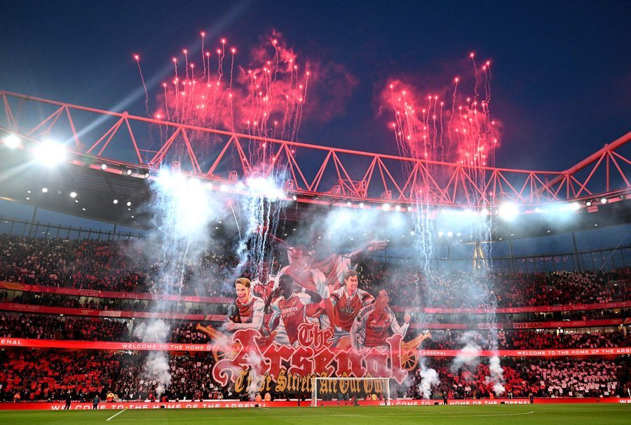 Arsenal's Emirates Stadium at dusk, red flares rising above a tifo banner of club legends draped across the stands