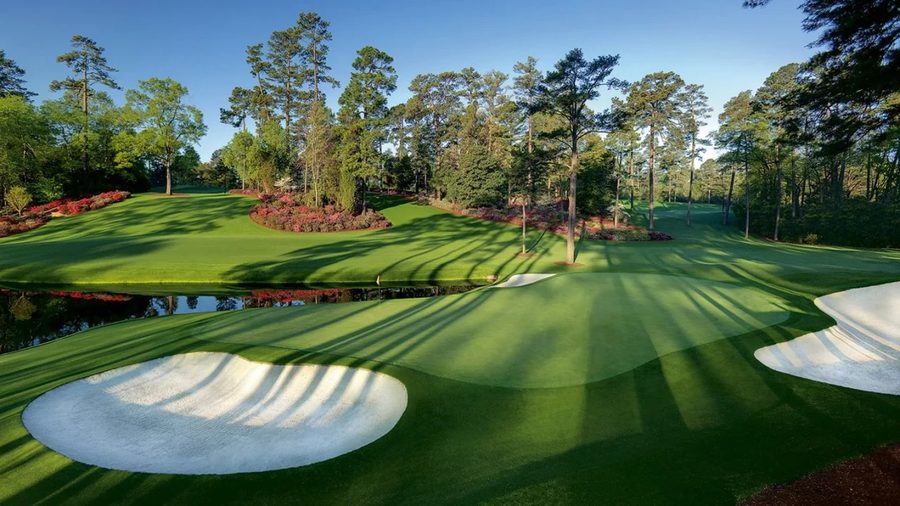 A sunlit golf hole with white sand bunkers and a water hazard reflecting pink azalea blooms