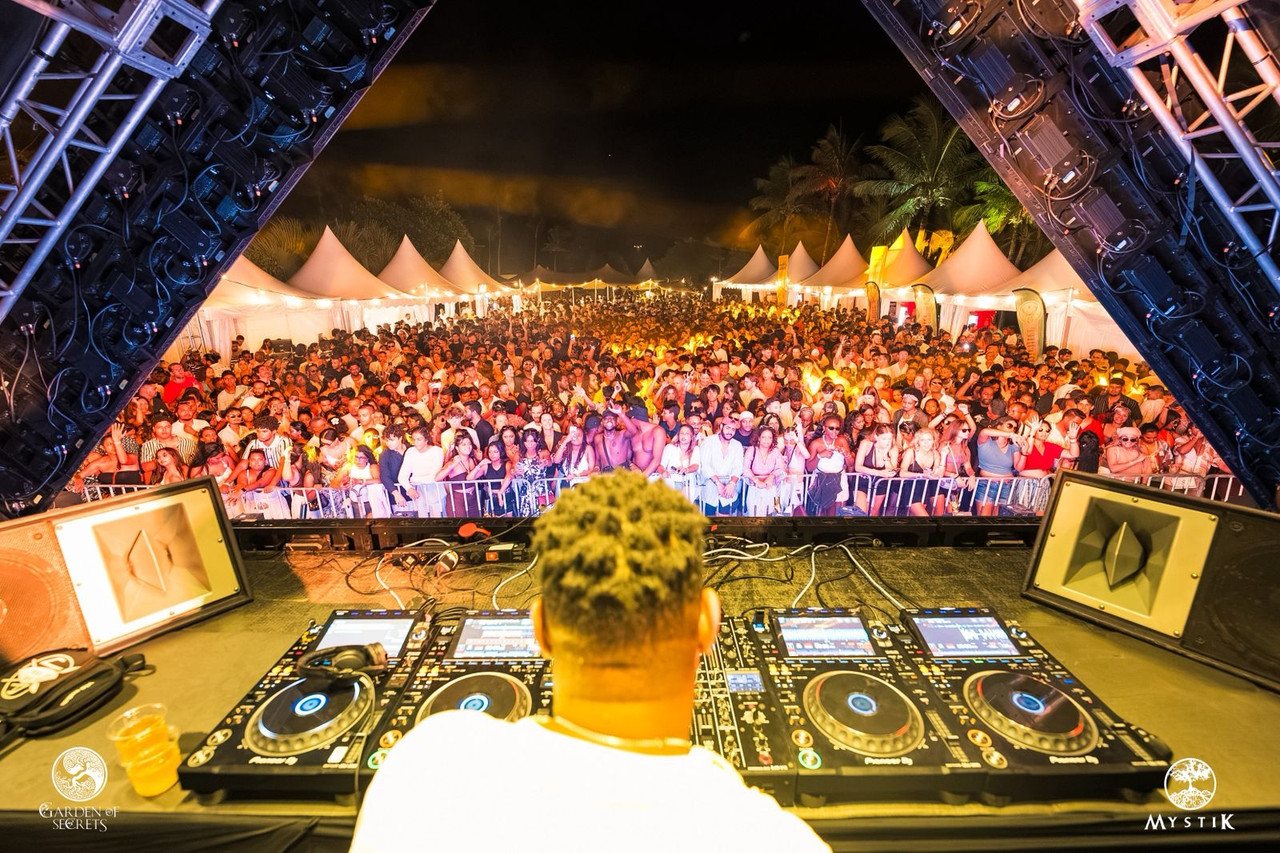 Backstage view from behind the DJ console, looking out over a crowd pressed against the front barrier under the stage truss