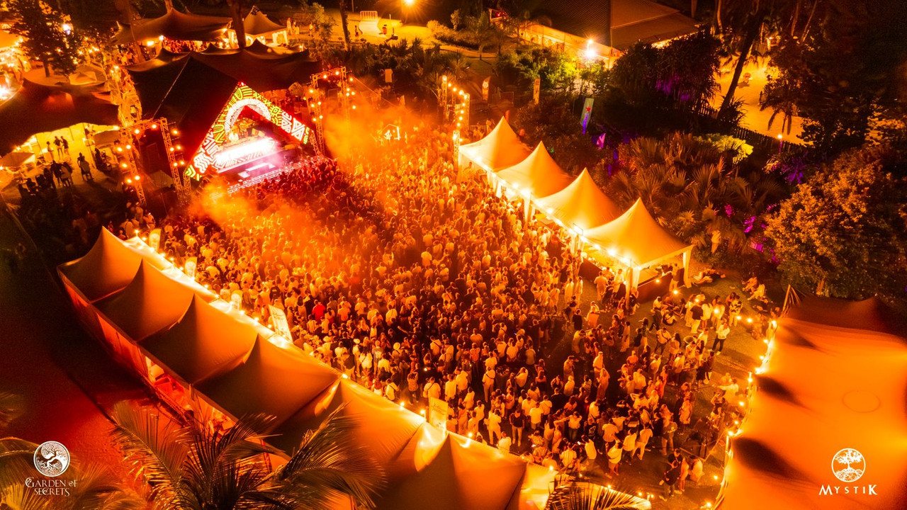 Drone view over the festival at night, the lit stage and dense crowd framed by rows of illuminated tents and palm trees