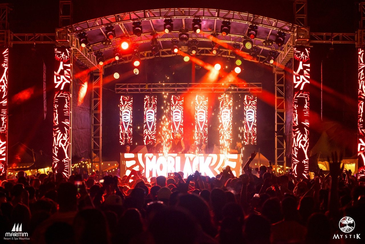 Main stage at night with an arched truss roof, vertical LED banners and cold-spark pyrotechnics firing from the DJ booth